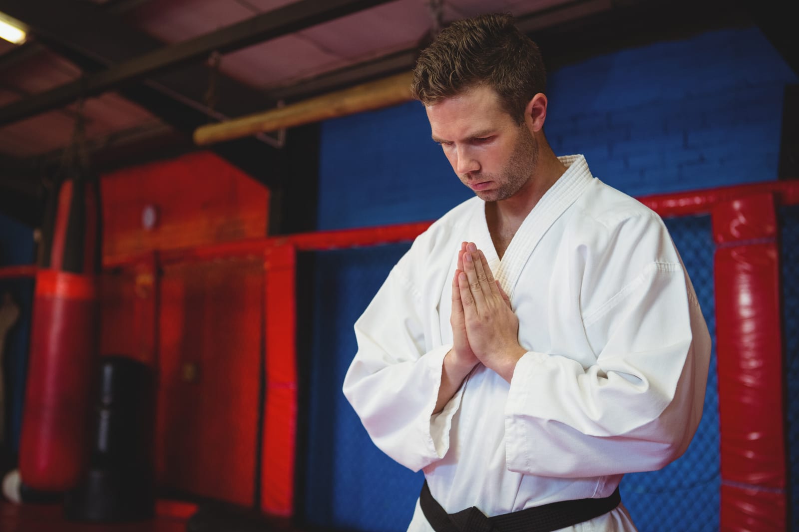A man in a white karate gi and black belt bows with palms pressed together inside a training gym.