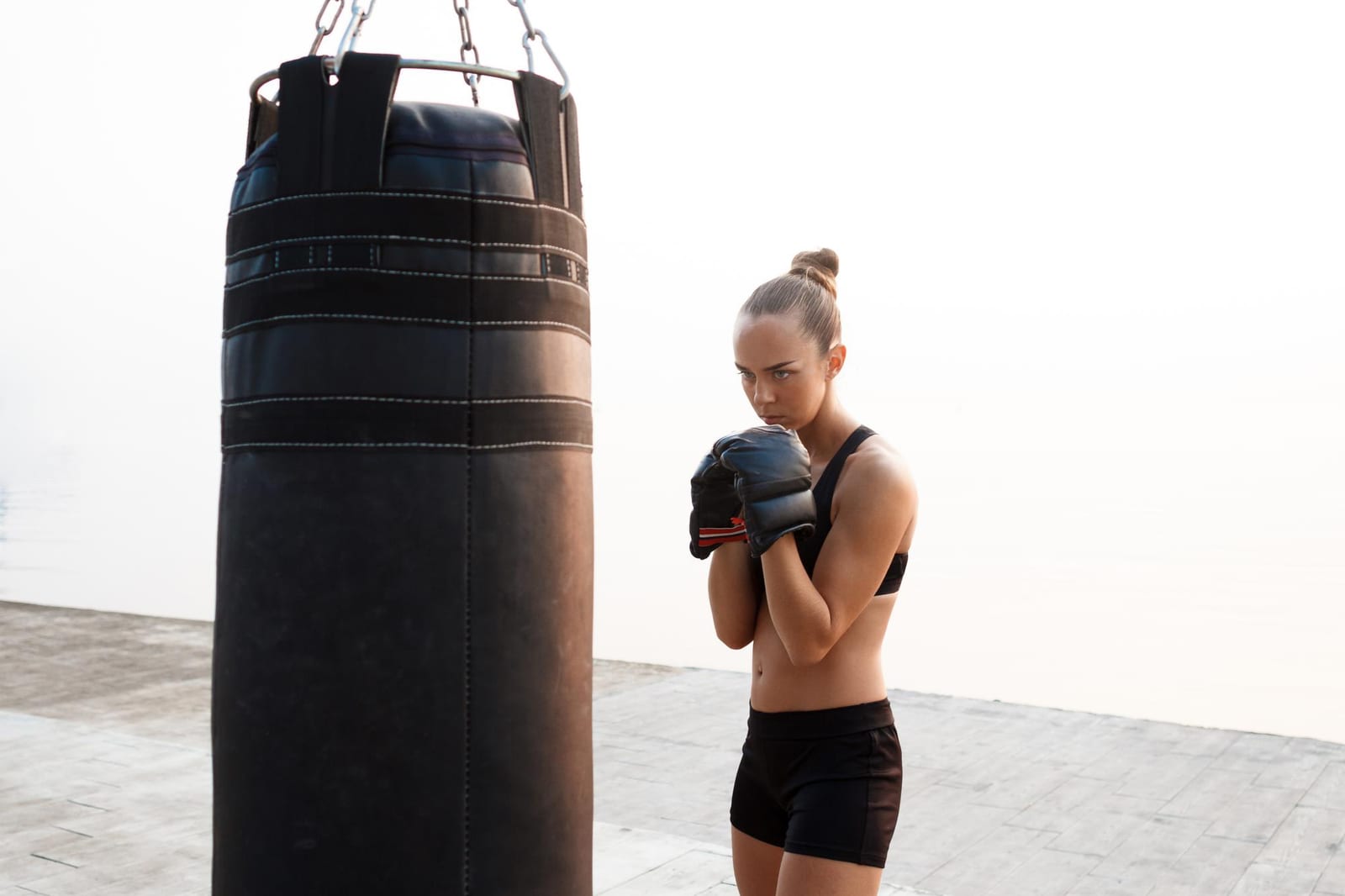 A woman in black sportswear and gloves holds a boxing guard beside a hanging heavy bag.
