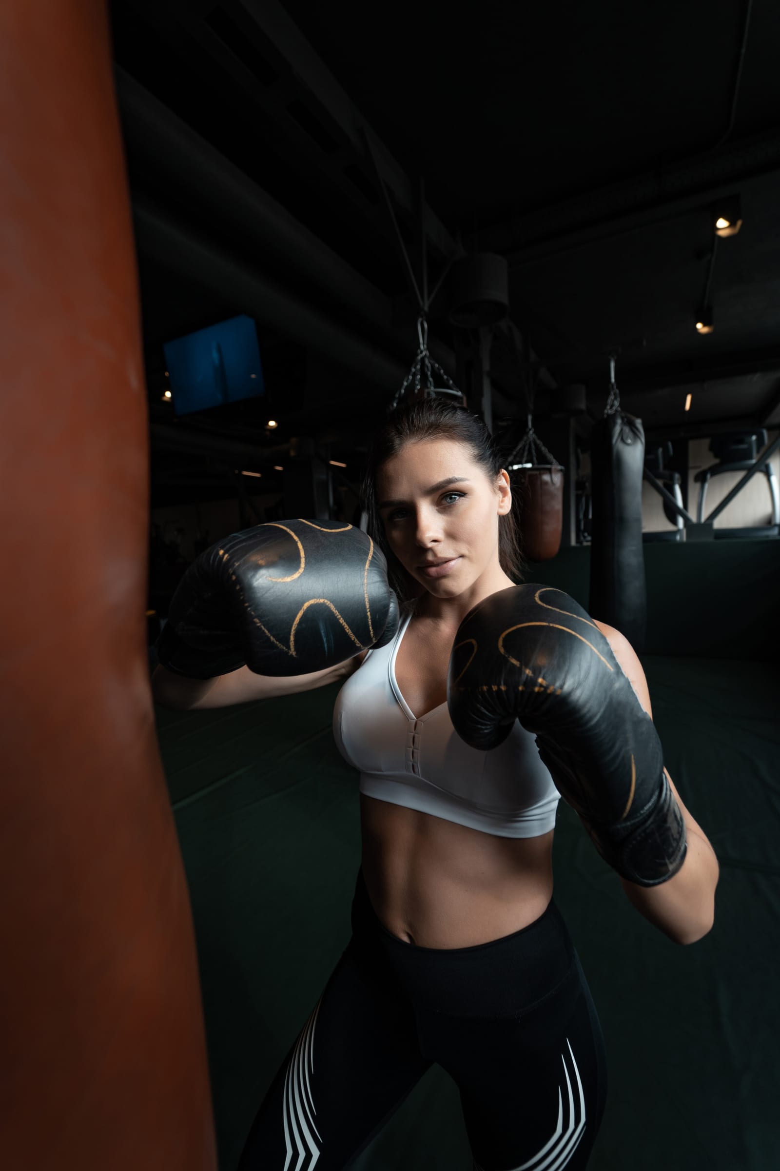 A woman in black boxing gloves holds her guard beside a heavy bag in a dark gym.