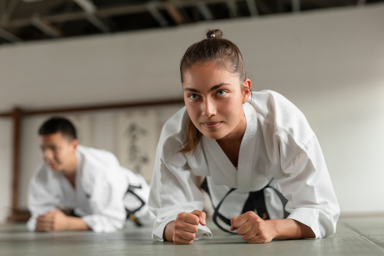 A woman in a white gi and black belt holds a low fist position on the dojo mat beside a fellow student.