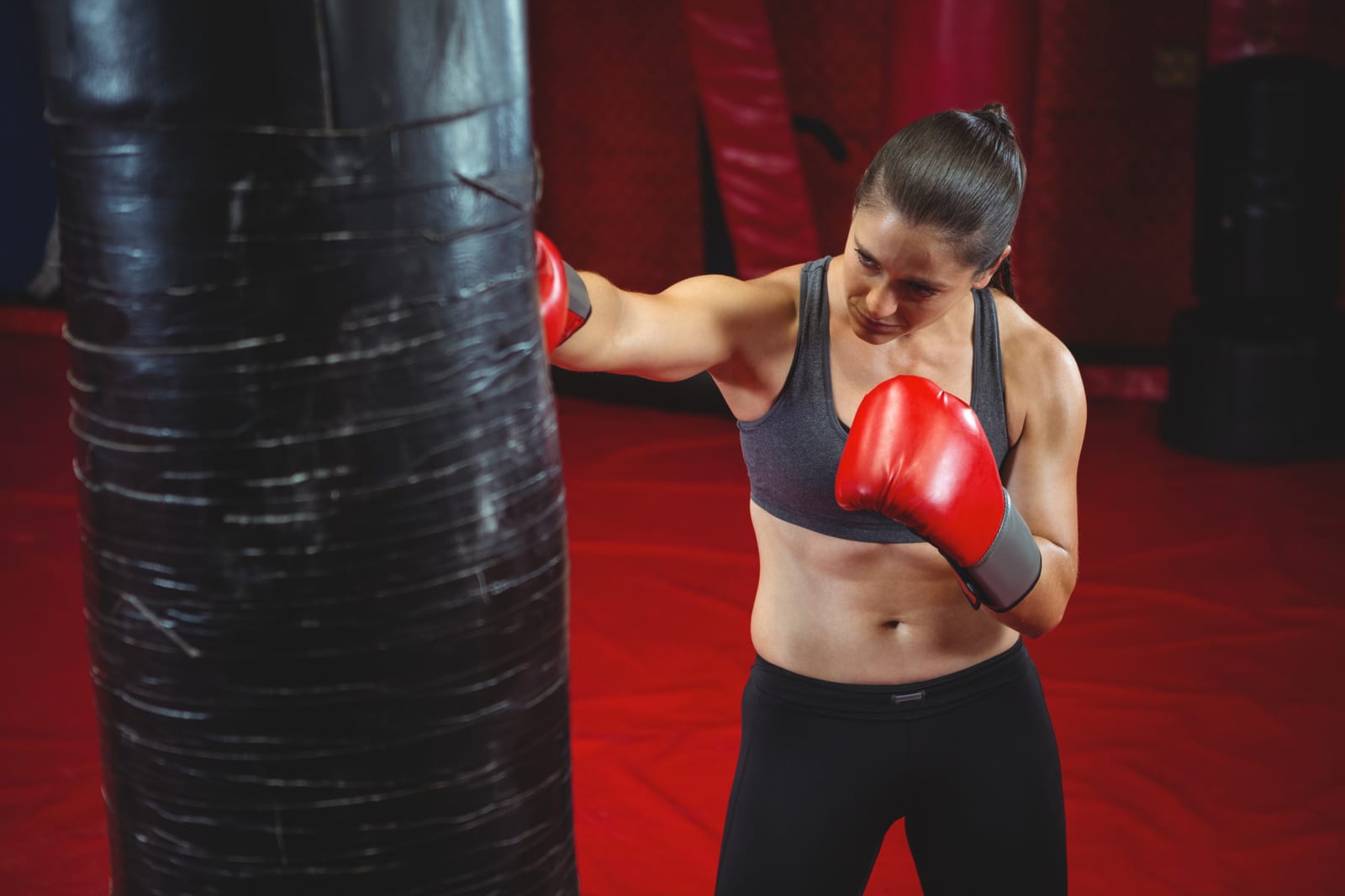 A woman in red boxing gloves punches a heavy bag in a red-walled gym.