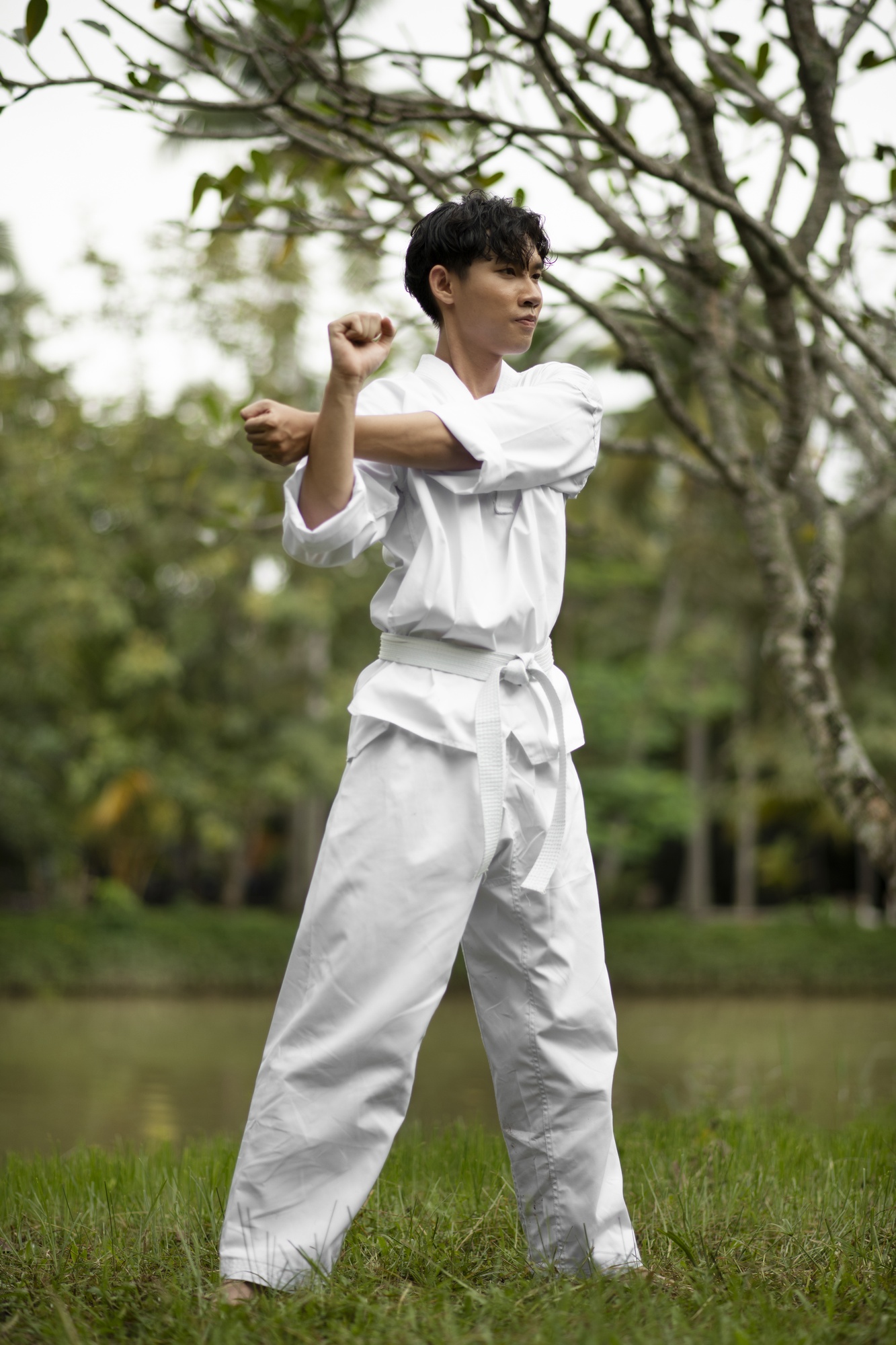 A martial artist in a white gi performs a blocking stance on grass beside a river.