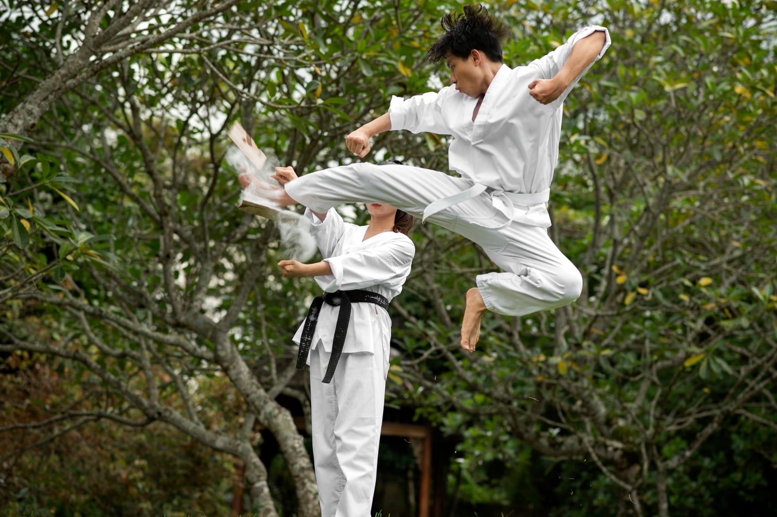 A martial artist in a white gi launches a flying kick through a board held by a partner outdoors.