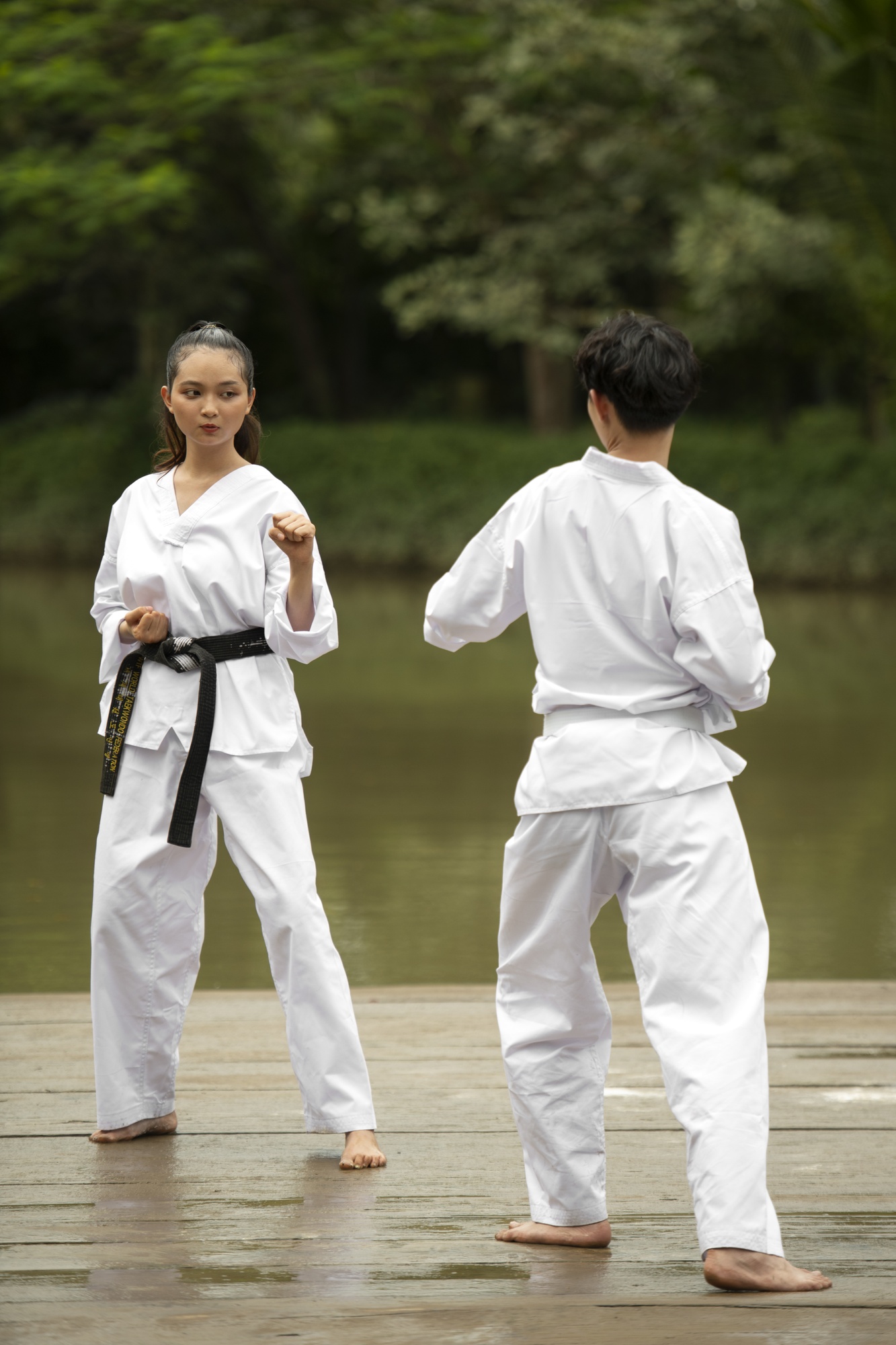 Two martial artists in white gis face each other in stance on a wooden deck by a river.